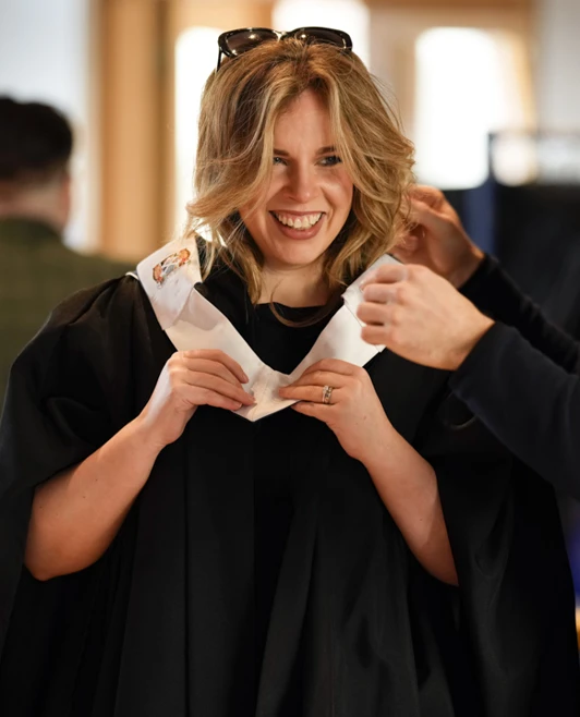 Smiling graduate receiving help with her graduation hood before the Glasgow Kelvin College 2025 ceremony. Smiling graduate receiving help with her graduation hood before the Glasgow Kelvin College 2025 ceremony.